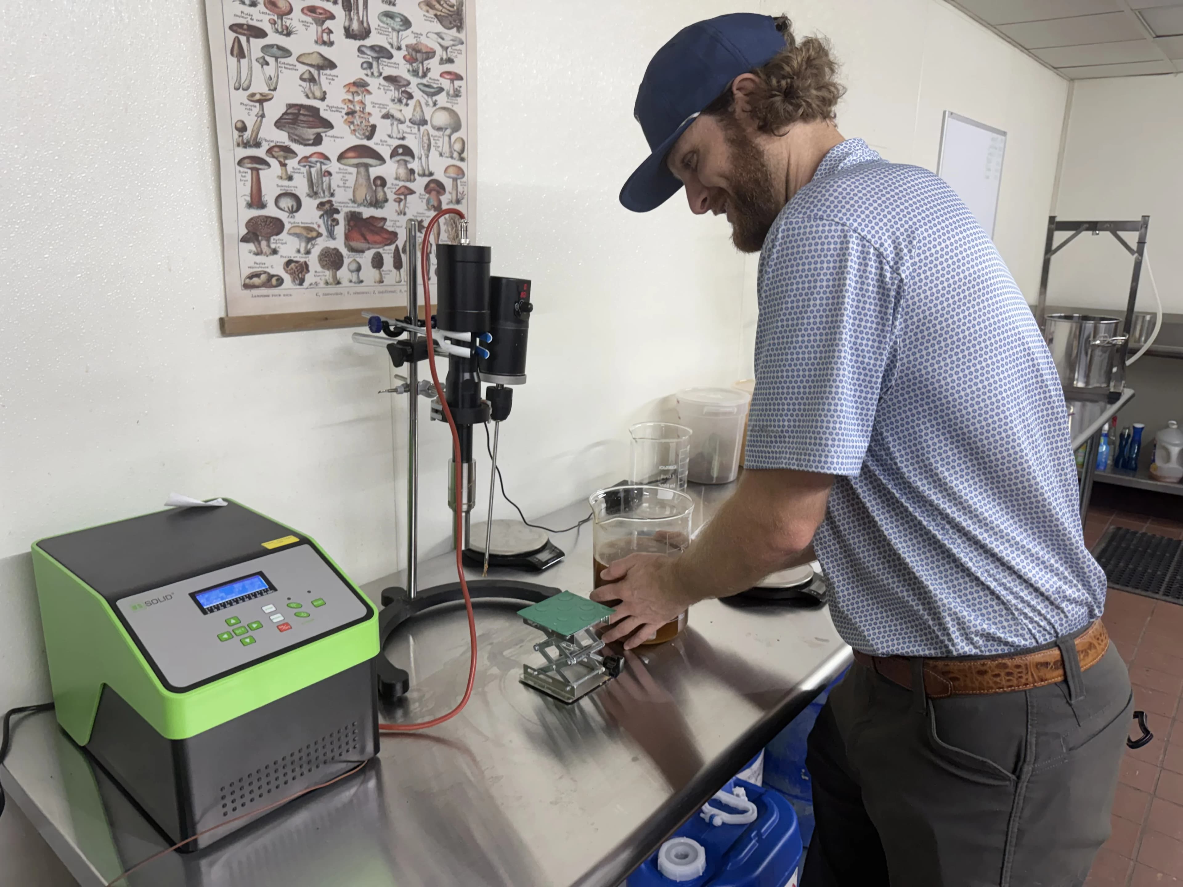 Samuel Sawyer working at a lab bench with extraction equipment and a mushroom identification poster on the wall behind him