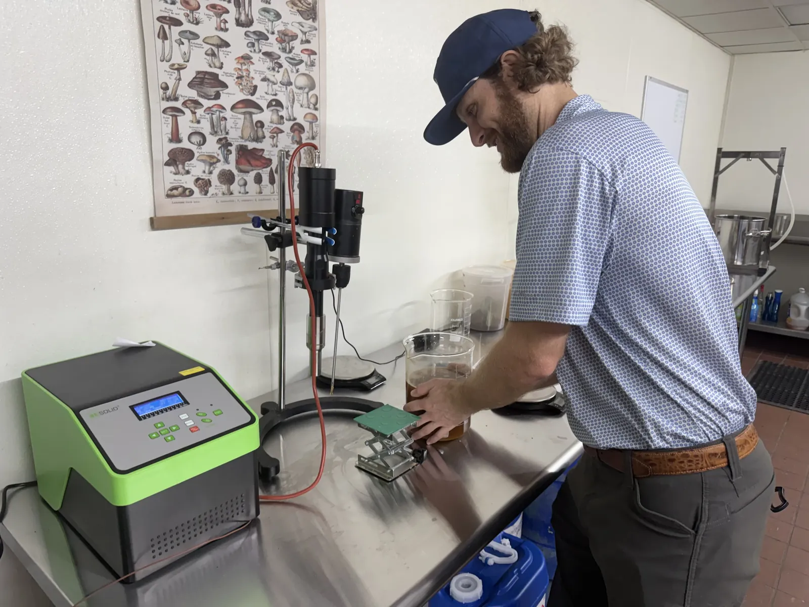 Samuel Sawyer working at a lab bench with extraction equipment and a mushroom identification poster on the wall behind him