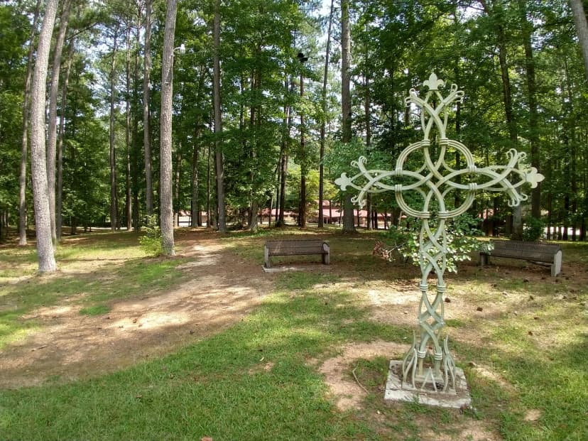 A weathered green Celtic cross standing in a pine grove at Camp Sumatanga, two empty wooden benches nearby, dappled summer light on the ground
