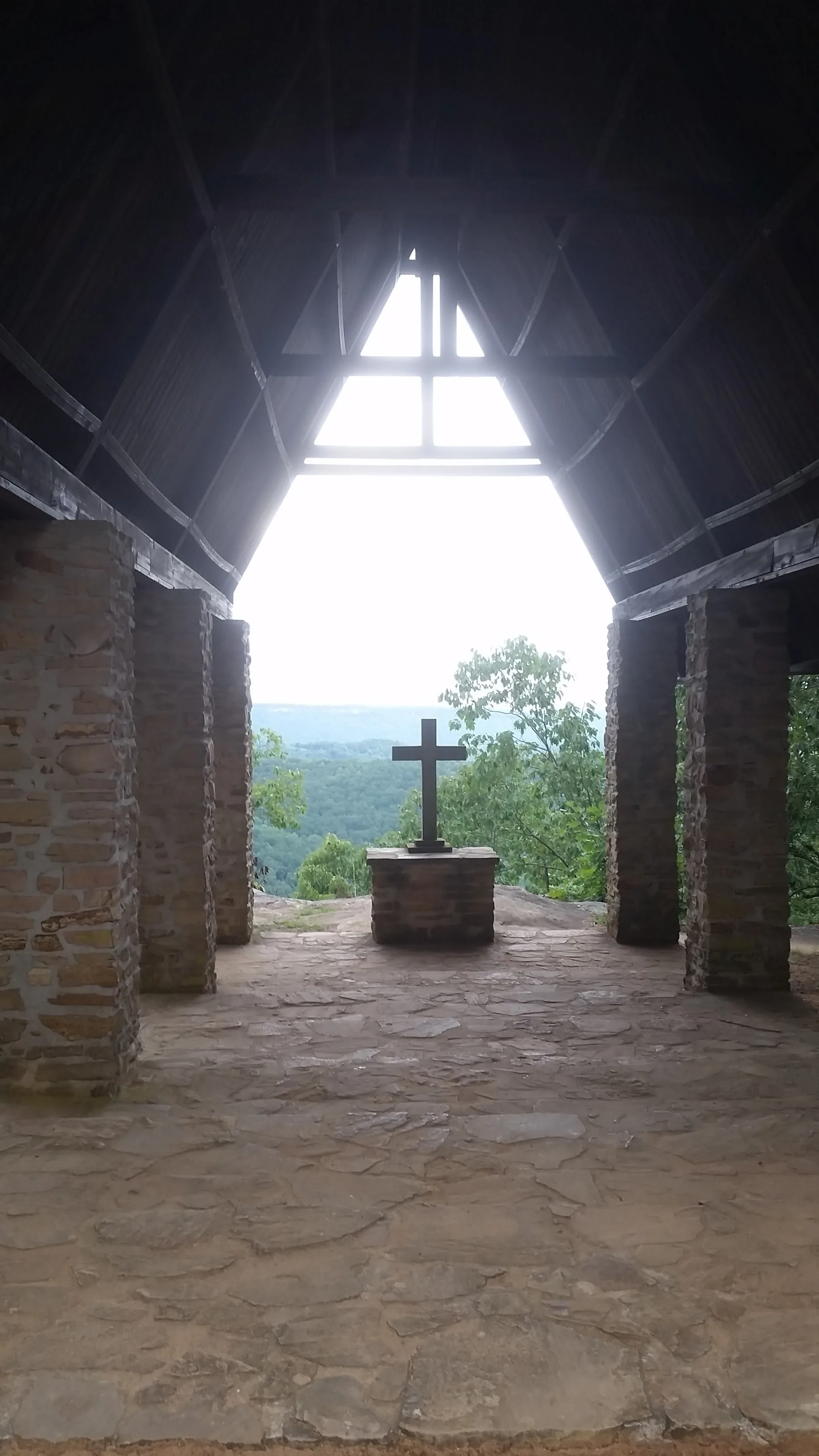 Interior of the open-air Chapel on the Mountain at Camp Sumatanga, a stone cross on a pedestal framed by the open triangular gable, the St. Clair County valley falling away beyond it
