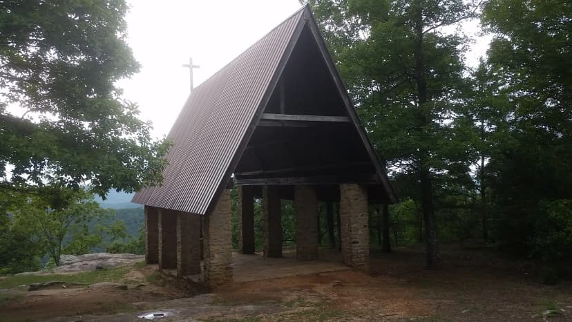 The open-air Chapel on the Mountain at Camp Sumatanga from outside, stone pillars, steep tin A-frame roof, a cross at the peak, standing on the ridge above the tree line