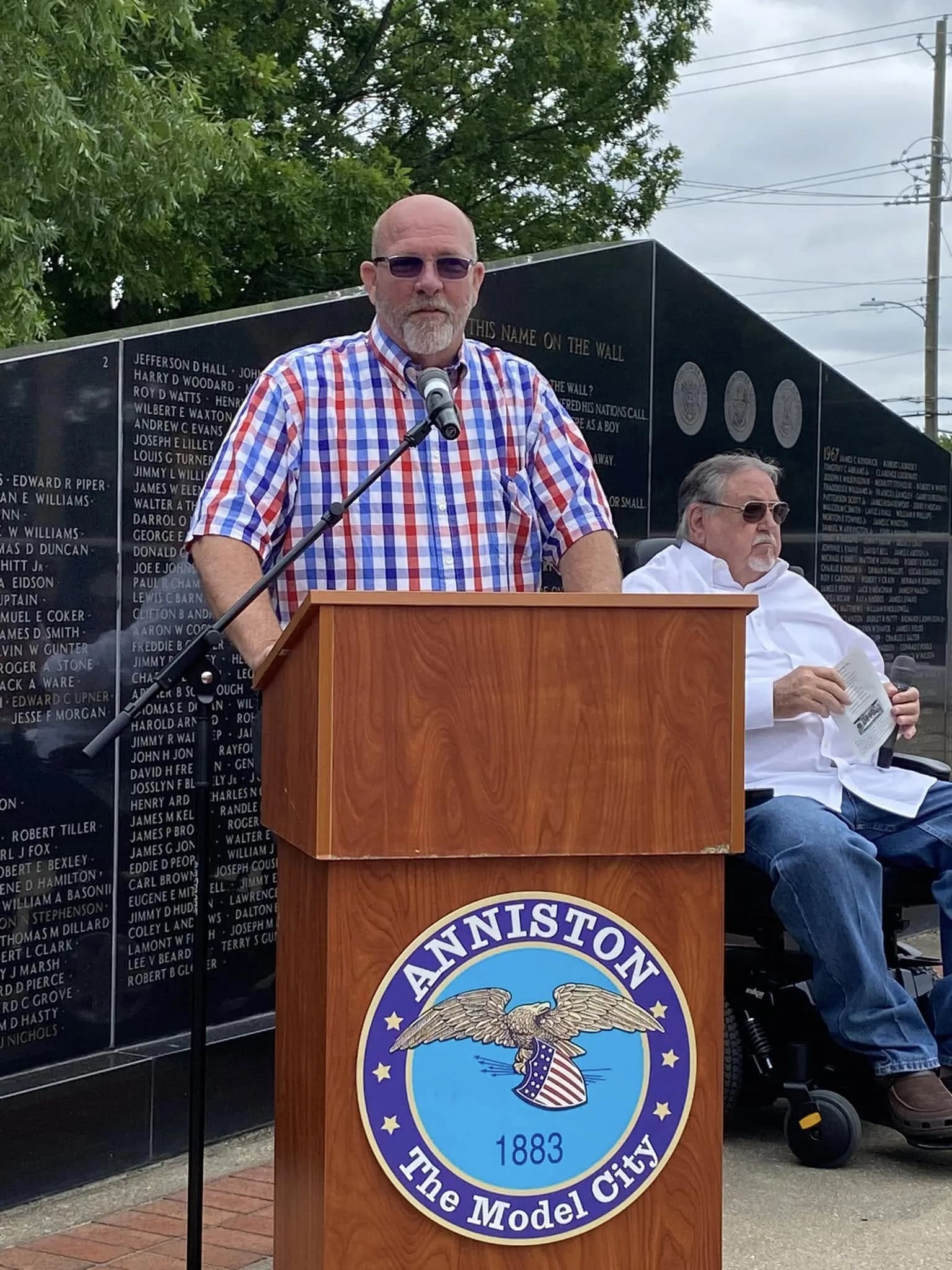 Jay Jenkins speaking at the Anniston Veterans Memorial podium, names on the memorial wall behind him, May 2023