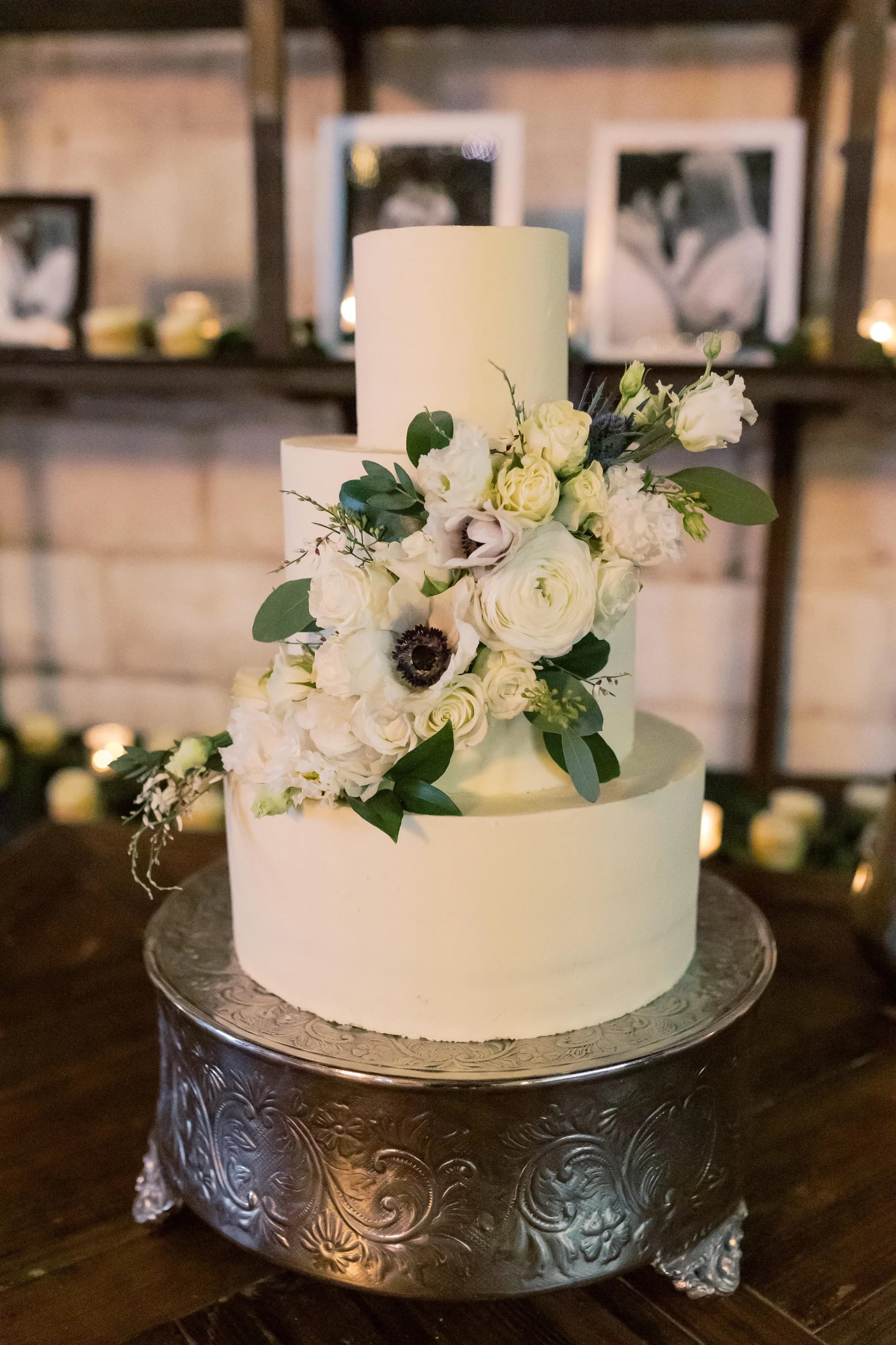 Three-tier white wedding cake adorned with white ranunculus, anemones, and eucalyptus greenery on a silver stand