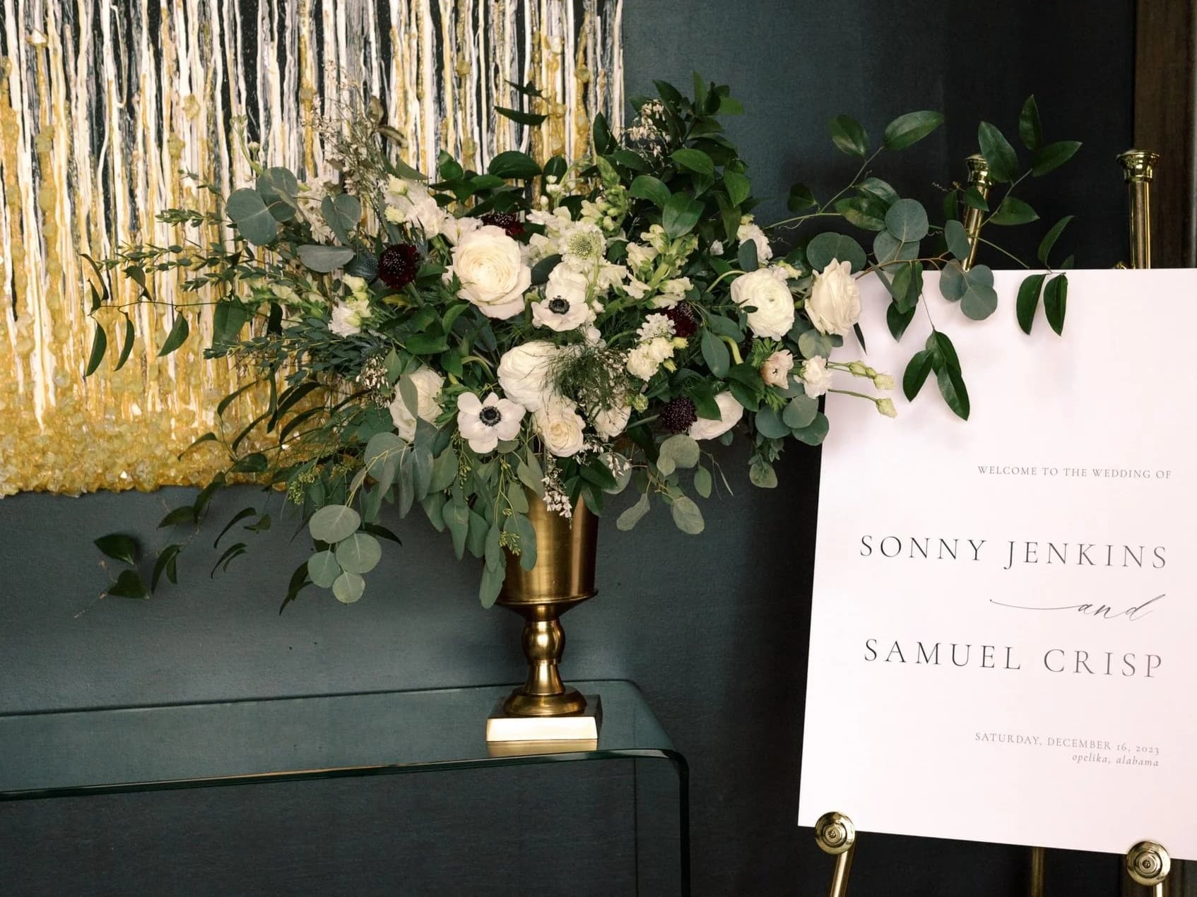 Entry display at the Jenkins-Crisp wedding — a brass urn overflowing with white ranunculus, anemones, and eucalyptus beside a welcome sign reading 'Sonny Jenkins and Samuel Crisp'
