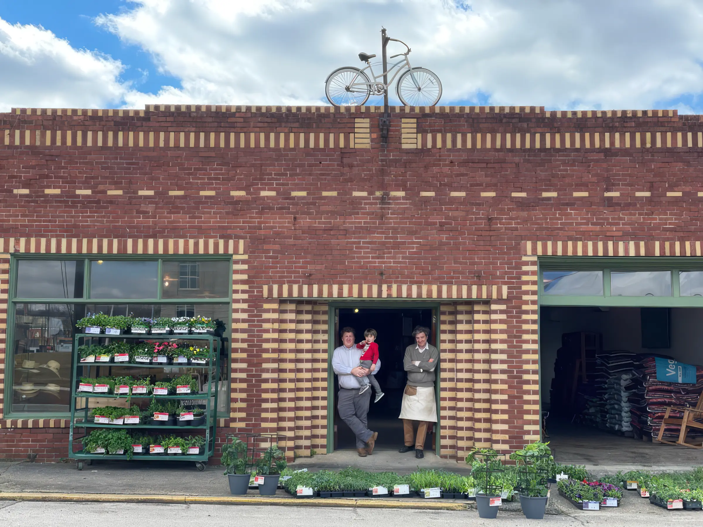 Lewis Downing and his father R.D. standing in the doorway of Downing and Sons on Gurnee Avenue in Anniston, with plants displayed out front and a bicycle mounted above the entrance