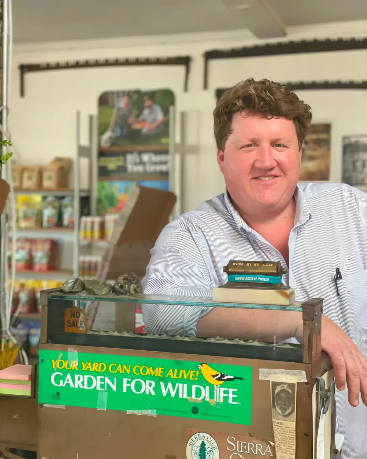 Lewis Downing leaning on the glass counter at Downing and Sons, with garden and seed products on shelves behind him